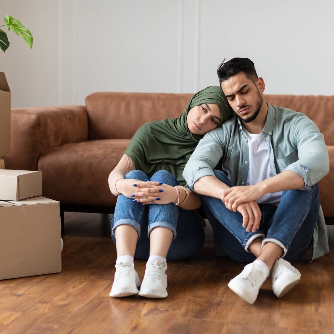 A couple sitting among moving boxes during a time of housing stress, leaning on each other