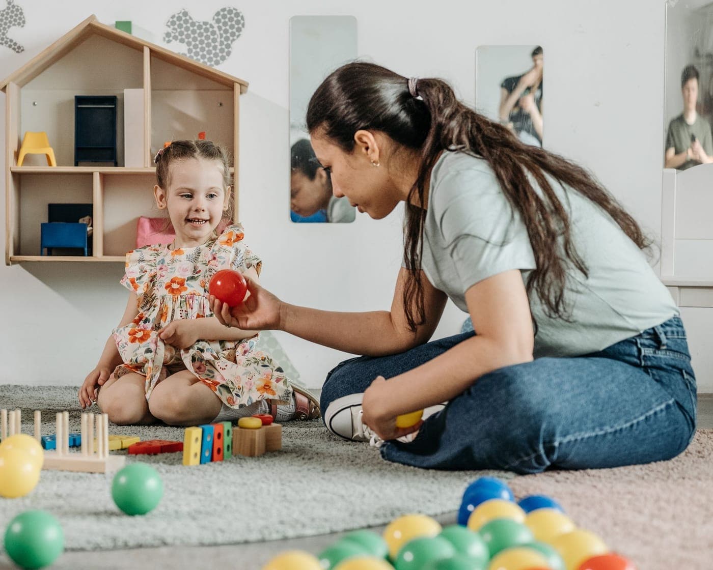 A practitioner sitting on the floor of a playroom passing a red ball to a smiling young girl
