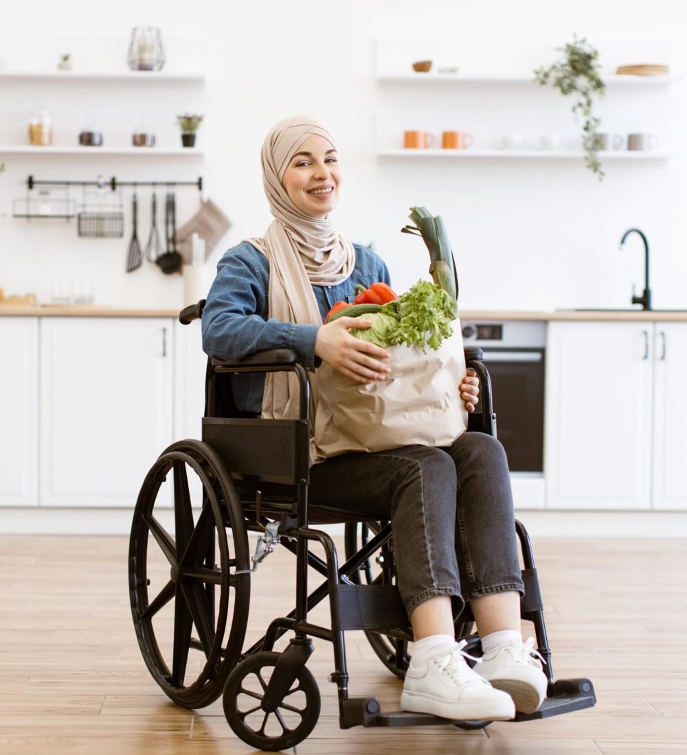 household-tasks-and-domestic-assistance-2048×2048 Muslim woman in wheelchair holding paper bag with fresh vegetables on background of modern kitchen. Smiling and enjoying healthy lifestyle and independence, showcasing inclusion and diversity.